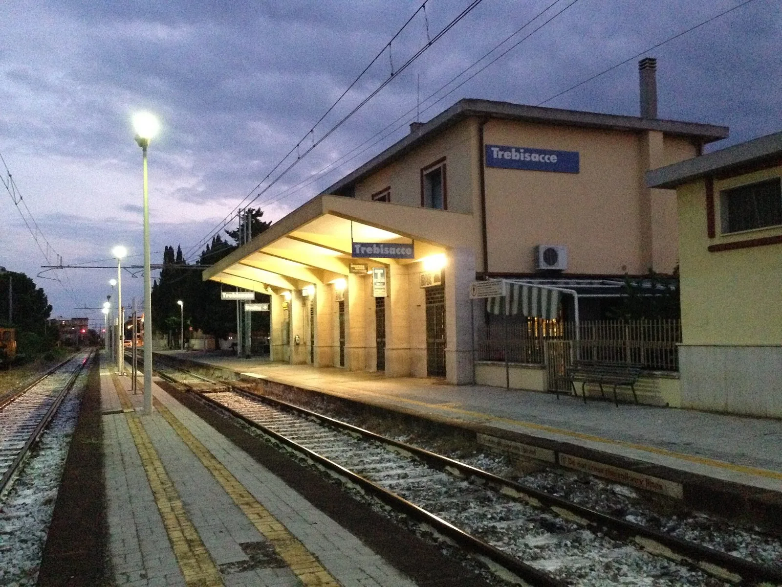 Il binario e la pensilina illuminata della stazione ferroviaria di Trebisacce al crepuscolo, con il cartello blu del nome della stazione