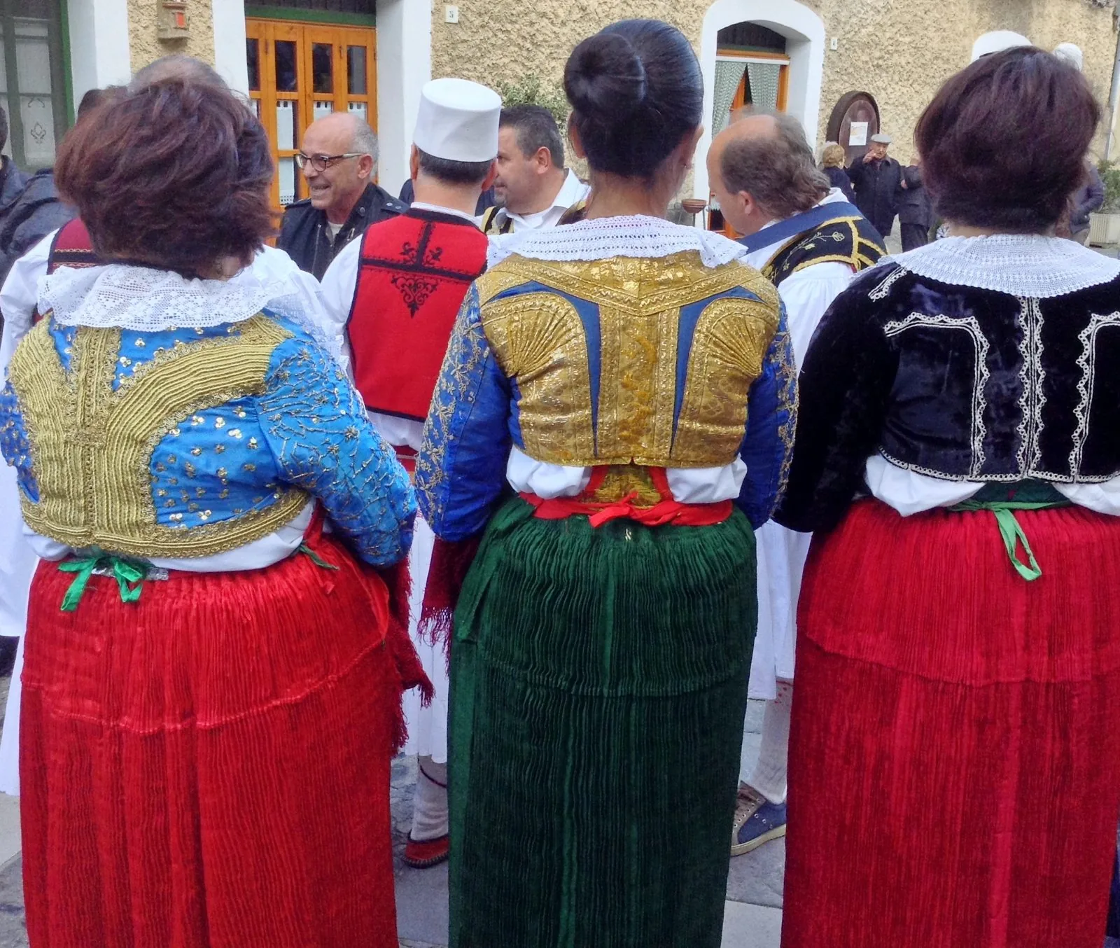 Donne di spalle in costume tradizionale arbëreshë a Civita, con corpetti ricamati d'oro, colletti di pizzo e gonne plissettate rosse e verdi, durante la festa delle Vallje