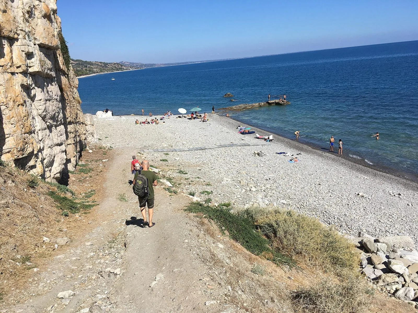 Spiaggia di ciottoli chiari sotto una parete di roccia, con bagnanti in riva e il mare Ionio che si apre verso la costa